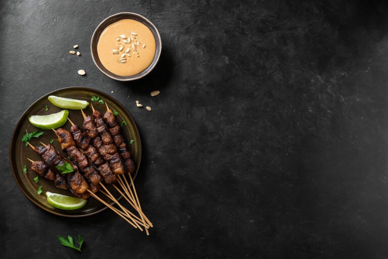 Beef satay skewers on a dark background with lime wedges and peanut dipping sauce